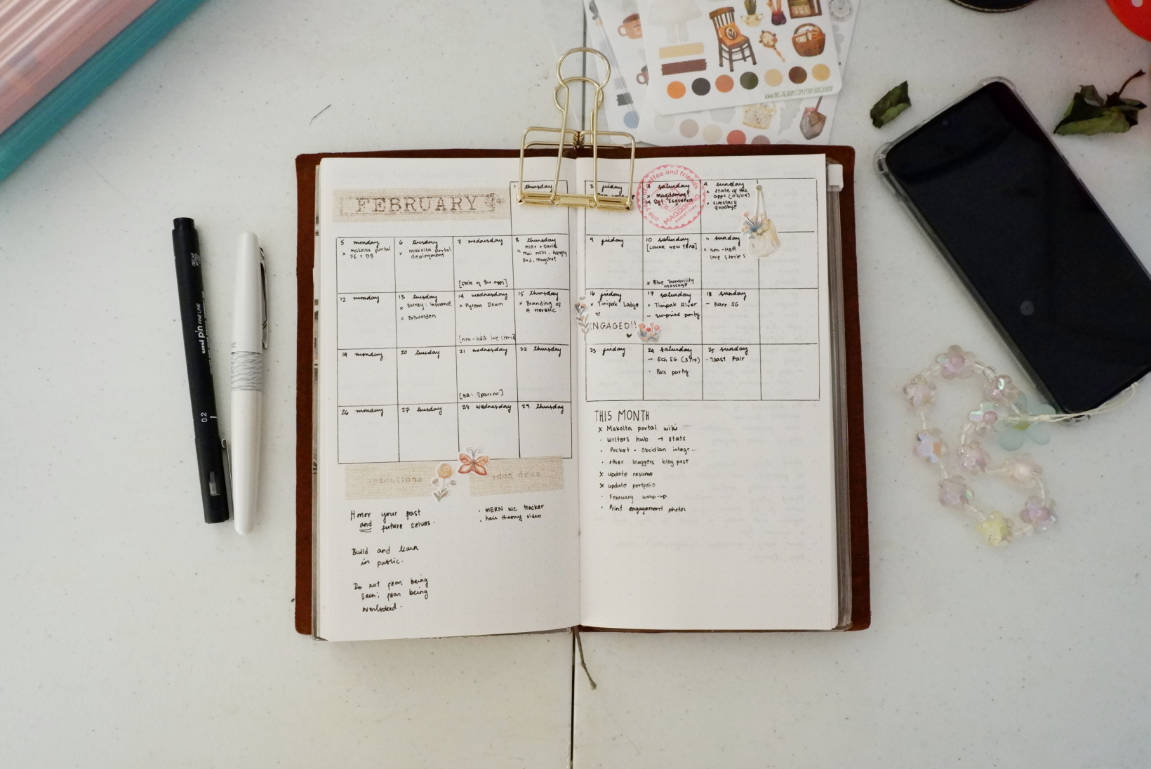 Flatlay image of an journal open to a February 2024 calendar spread. The notebook is surrounded by a pile of stickers on top, a white and a black pen to the left, and a phone with translucent charms to the right.